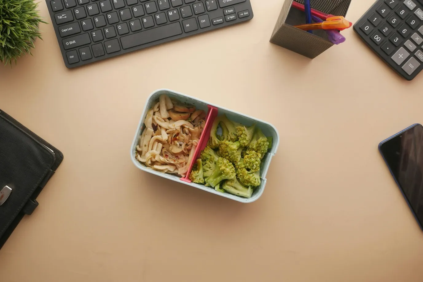 Healthy packed lunch with noodles and broccoli on an office desk beside a keyboard and phone