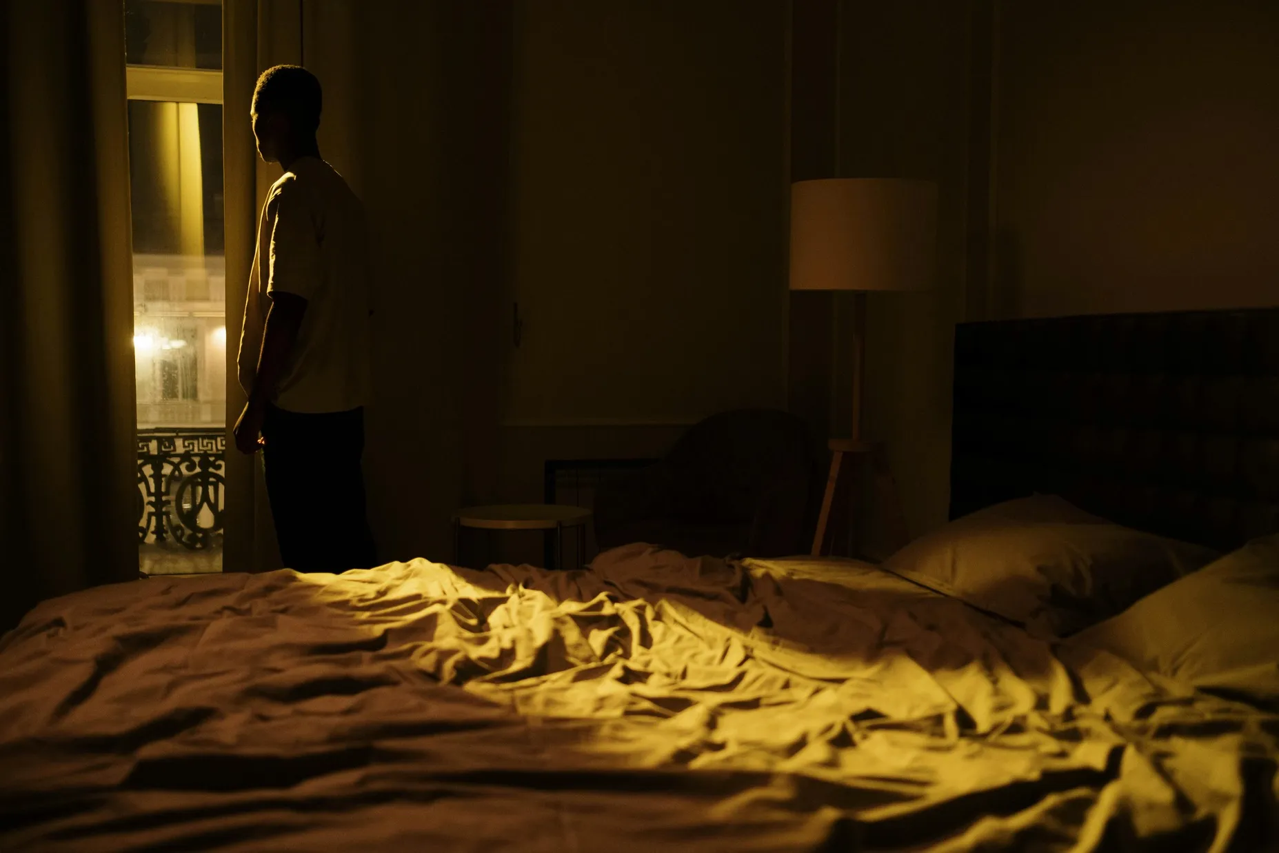 Person standing awake in a dim bedroom, while closing the curtains, beside an unmade bed at night.