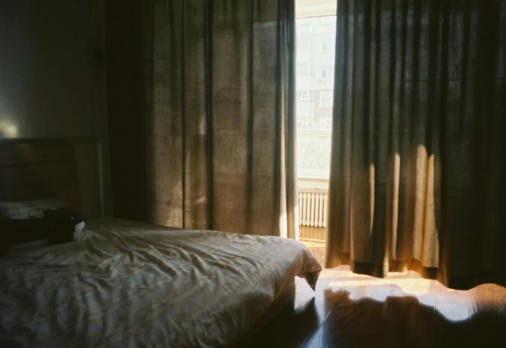 Bedroom with dark curtains partly open letting sunlight in near a bed