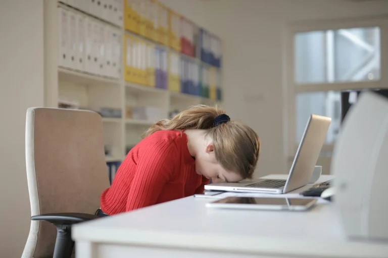 Busy professional resting her head on a desk beside a laptop in an office