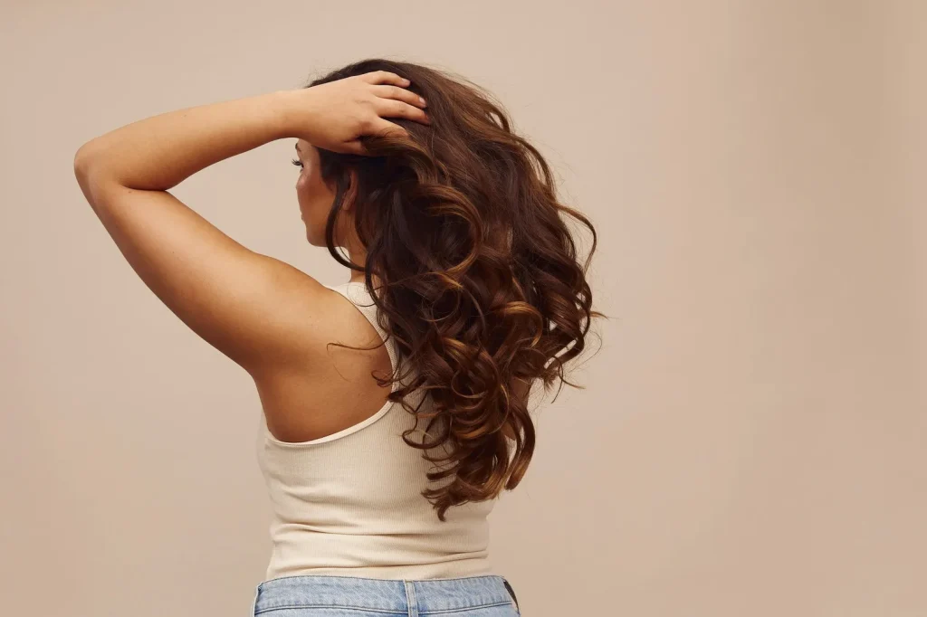 Woman seen from the back with long, glossy, softly defined wavy hair against a neutral background.