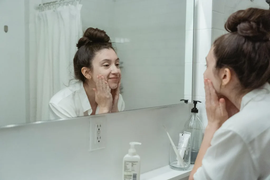 Woman applying facial cleanser in front of a bathroom mirror during a summer skin care routine for oily skin.